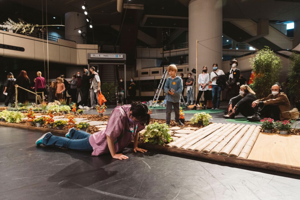 Woman leaning on the ground in the foreground. In the background there are many people in a large area. A footbridge with plants is set up next to the woman