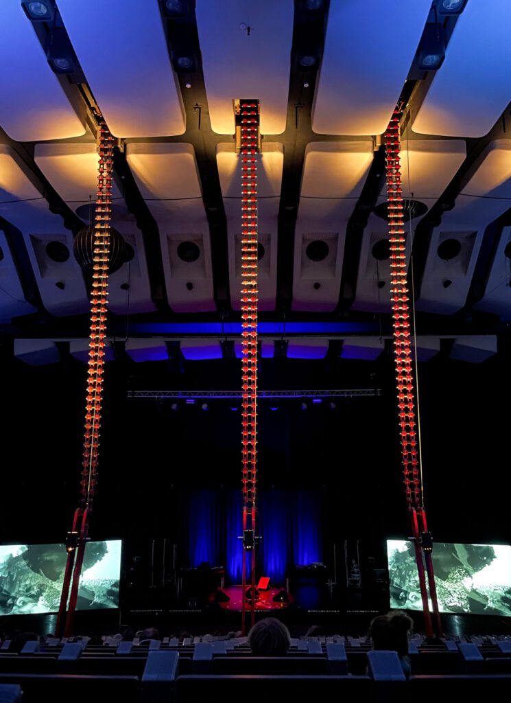 Metal chains connected to rows of chairs. A stage with screens can be seen on the floor in the background