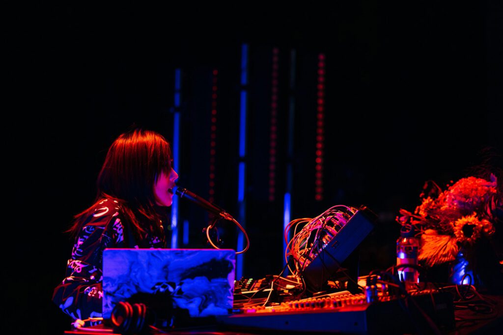 Woman singing into a microphone. There is a lot of technology in front of her and the stage is colorfully lit