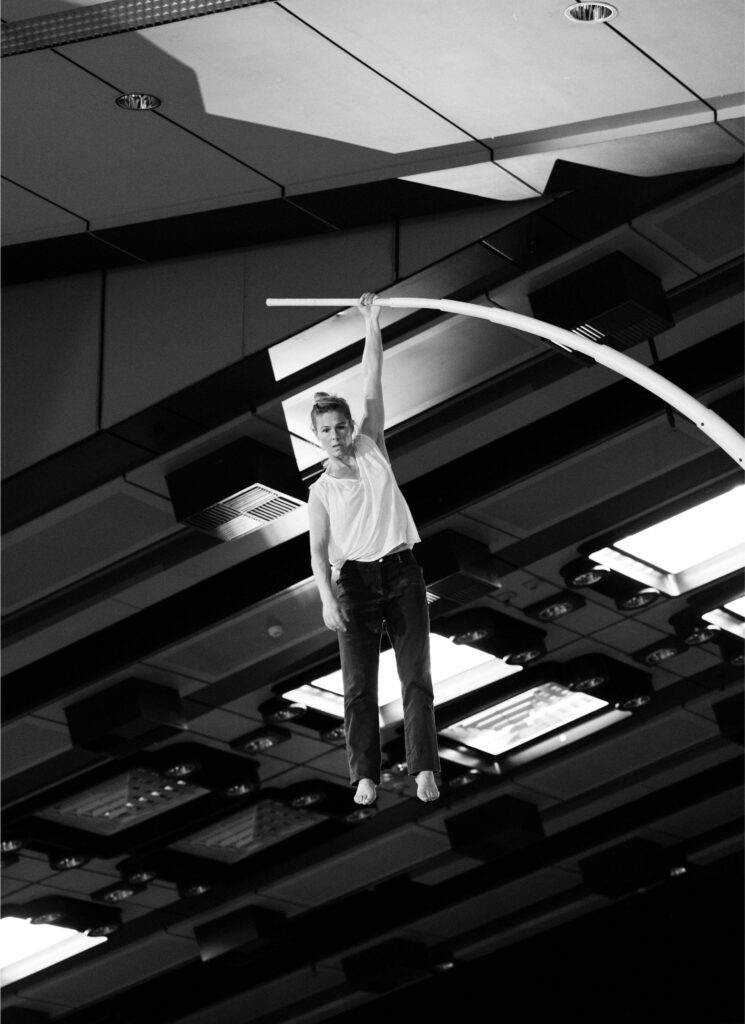 Woman hangs with one arm on a bent pole with a neutral expression on her face. A room with a ceiling can be seen in the background. Black and white photo