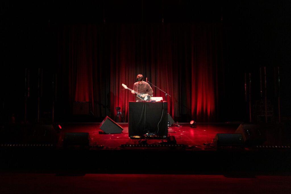 Man with guitar on a small stage in a dark room with red lighting