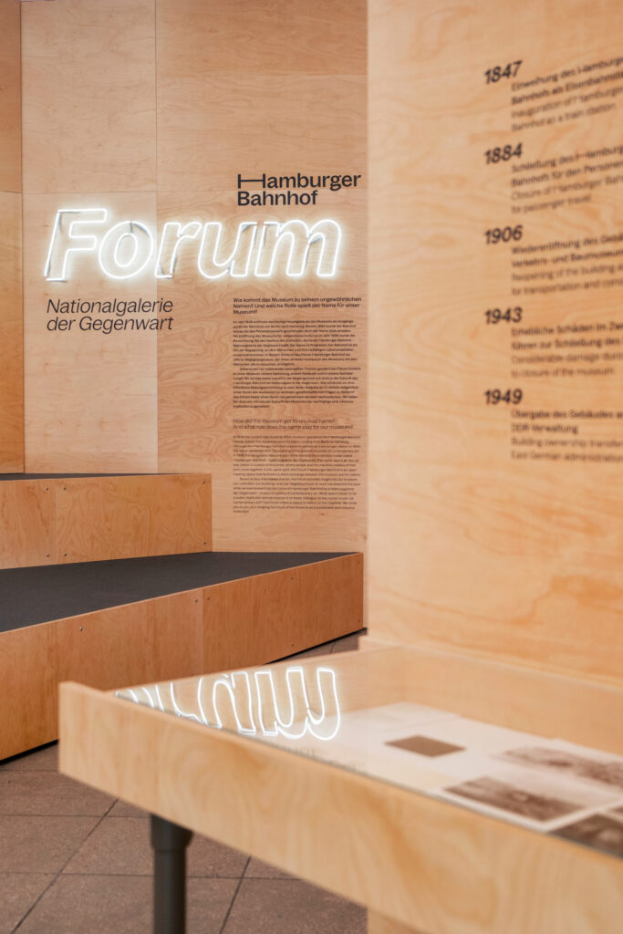 Wooden room with glowing lettering in the background. Small black lettering with dates and text in the foreground above exhibition boxes