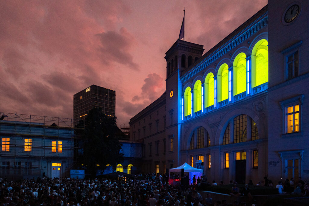 Große Veranstaltung vor dem Museum. Rosafarbener Abendhimmel mit beleuchteten Bögen des Museums.