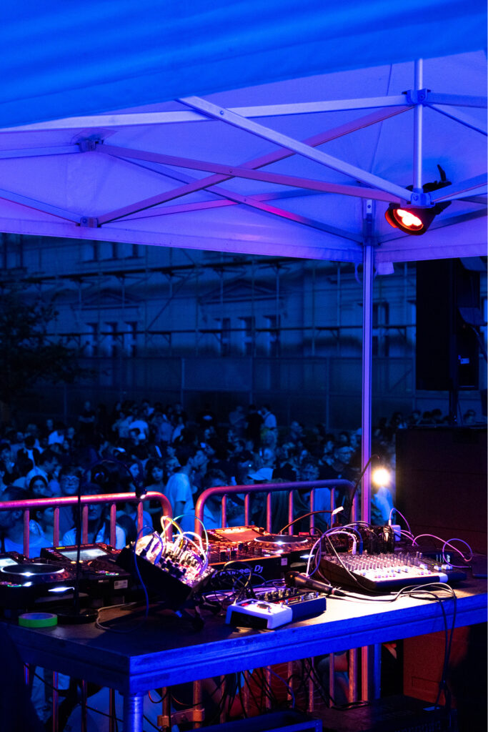 DJ set in the foreground with lots of people in the background. At night with blue light