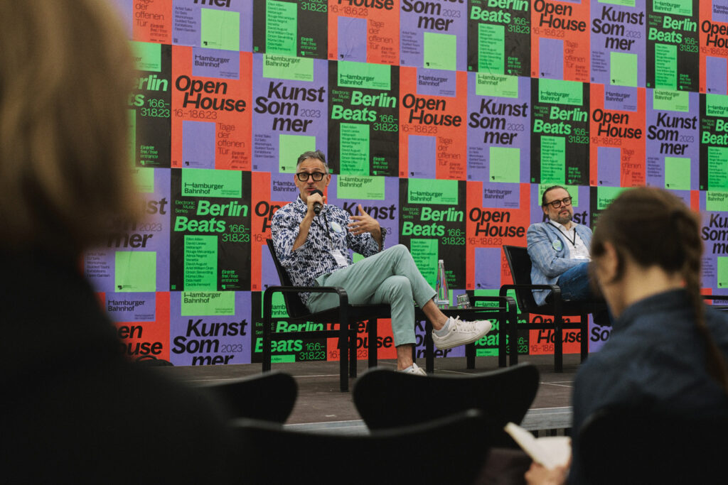 Two men sit in chairs on a stage and give a lecture. In the background, the entire wall is covered with posters. These are colorful and have large lettering and spaces